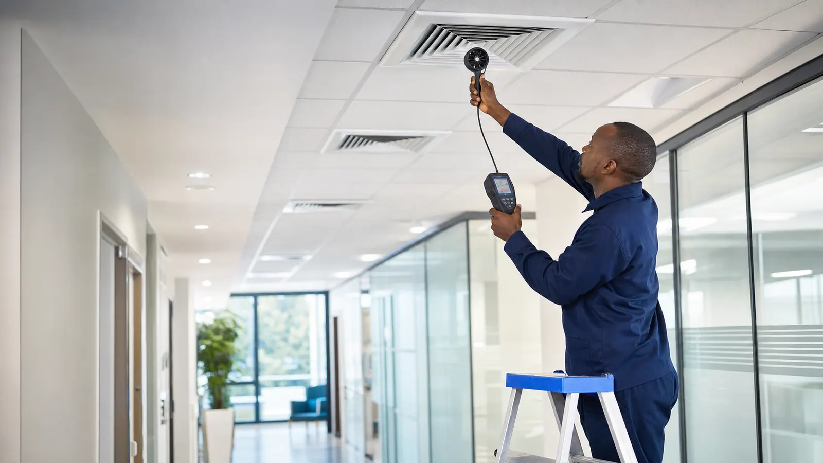 Technician measuring airflow at a ceiling diffuser in a commercial office corridor